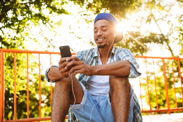 公園でスケートのランプに携帯電話で音楽を聴くイヤホンで幸せな若いアフリカ男 — ストック写真