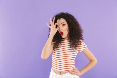 Ok işaret parmak Studio violet arka plan üzerinde izole delikten bakarken gösteren yaz aşınmaya kıvırcık saçlı Genç kadın fotoğrafı