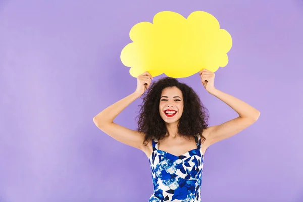 Image of charming happy woman with curly hair in dress smiling and holding blank bubble above her head with copyspace for your text isolated over violet background in studio