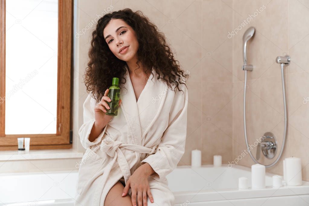 Photo of young pretty woman in bathroom holding shower gel shampoo in hands.