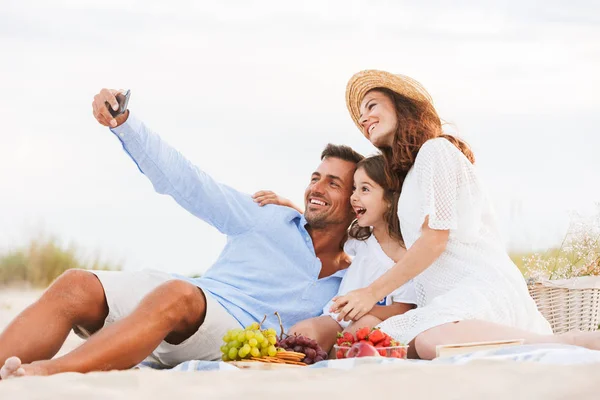 Joyful family with father, mother, daughter having picnic at the beach, taking selfie