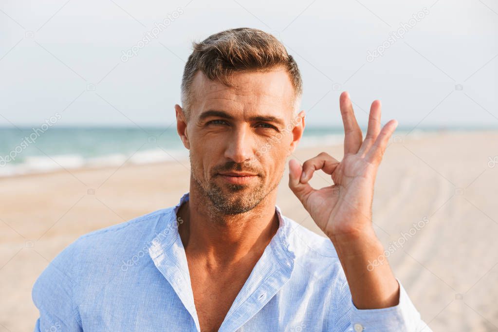 Portrait of a handsome man in shirt standing at the beach, showing ok gesture