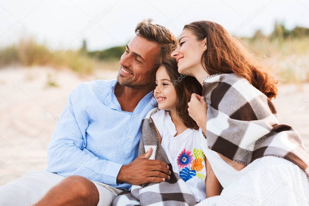 Image of young cute parents hugging outdoors at the beach with their daughter under plaid. Looking aside.
