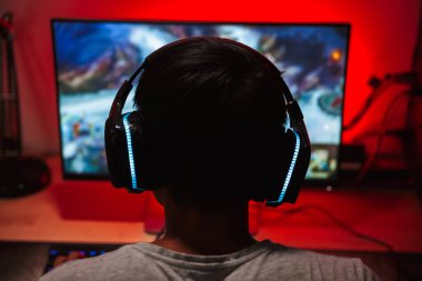 Portrait from back of young gamer guy looking at screen and playing video games on computer in dark room wearing headphones clipart