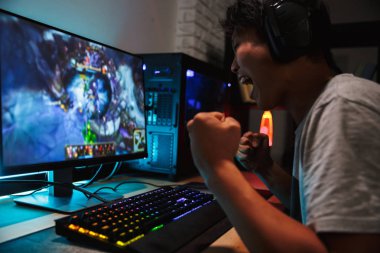 Joyful asian teen gamer man playing video games on computer in dark room wearing headphones and using backlit colorful keyboard clipart