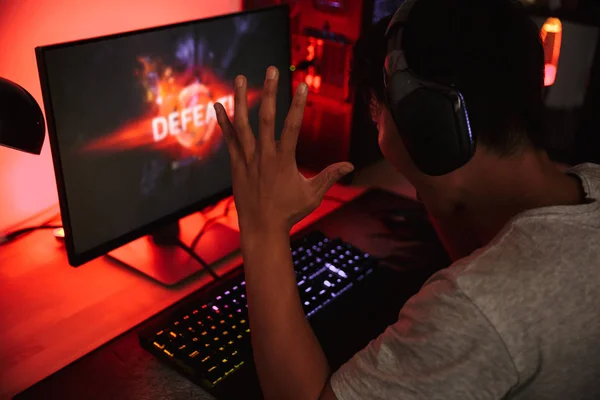 ビデオ ゲームをプレイしながら失う動揺してアジアのゲーマー少年の肖像ヘッドフォンを身に着けていると カラフルなキーボードのバックライトを使用して暗い部屋でコンピューターの — ストック写真