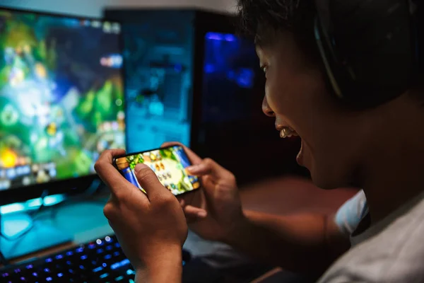 スマート フォンとヘッドフォンを身に着けていると カラフルなキーボードのバックライトを使用して暗い部屋でコンピューターのビデオゲームで遊ぶ幸せの 代のゲーマー少年の肖像画 — ストック写真