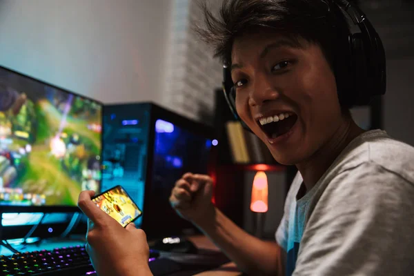 アジアの幸せゲーマー少年のビデオ ゲームをプレイしながら歓喜のスマート フォンとコンピューターの暗い部屋でヘッドホンをつけているとバックライト付きカラフルなキーボードを使用して — ストック写真