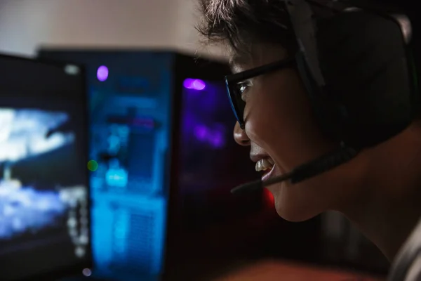 マイク付きヘッドフォンを着用し バックライト付きカラフルなキーボードを使用して暗い部屋でコンピューターのゲームをオンラインでビデオを再生アジアの満足しているゲーマー少年の写真のクローズ アップ — ストック写真