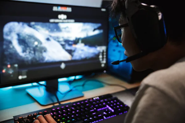 マイク付きヘッドフォンを着用し バックライト付きカラフルなキーボードを使用して暗い部屋でコンピューターのオンライン ビデオ ゲームを遊ぶゲーマーのアジアの少年の写真のクローズ アップ — ストック写真