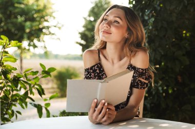 Café outdors kitap ile parkta oturan sevimli neşeli genç ve güzel kadın görüntüsü. Bir kenara arıyorsunuz.