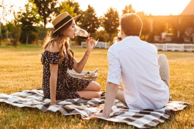 Outdors şarap içme gözlük tutan piknik üzerinde çıkarak oturan genç sevgi dolu çift.