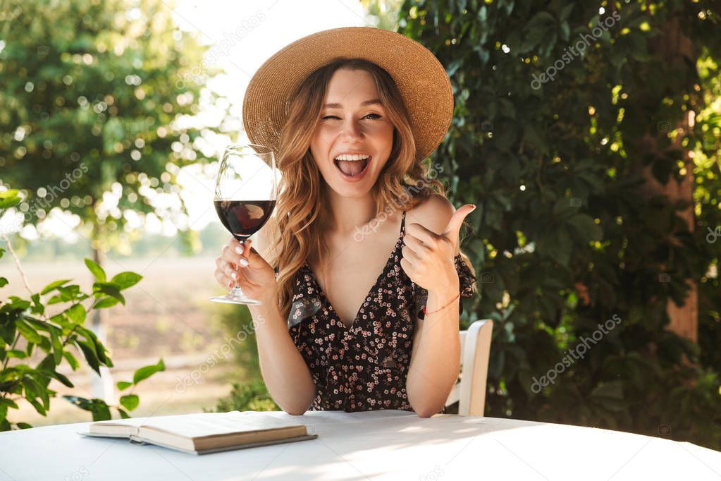 Image of cheerful lovely woman wearing straw hat drinking wine while sitting at table and reading book outdoor on nature