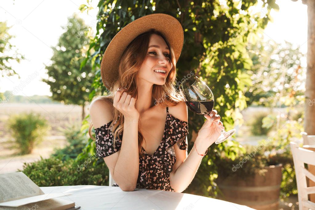 Image of cute pretty young woman sitting in cafe outdors in park with book holding glass drinking wine.