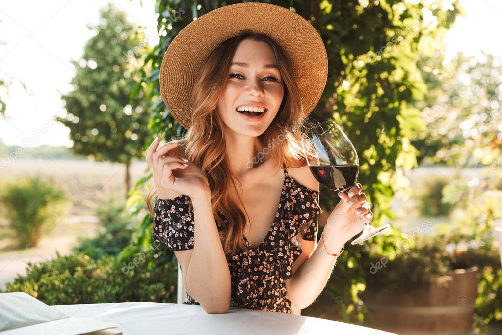Image of pretty young woman sitting in cafe outdors in park holding glass drinking wine.