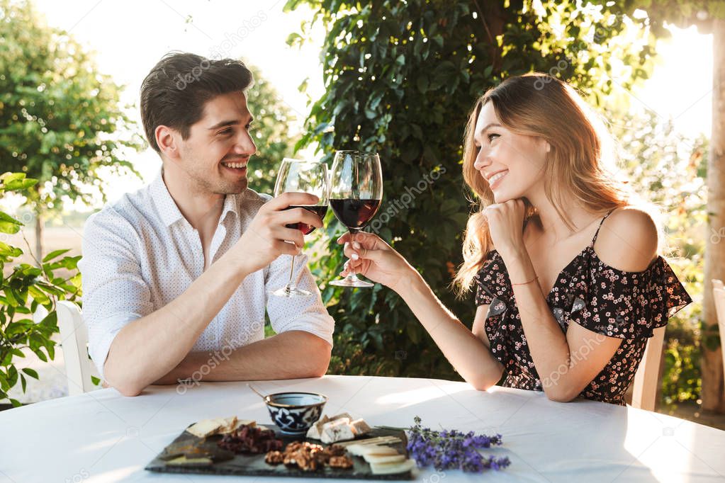 Picture of young loving couple sitting in cafe by dating outdors in park holding glasses of wine drinking.