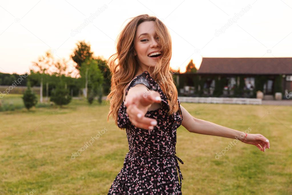 Photo of beautiful young woman 20s wearing dress smiling and giving hand to you while walking outdoor through green field in countryside