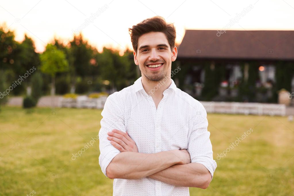 Photo of handsome young man wearing white shirt smiling while walking outdoor through green field in countryside