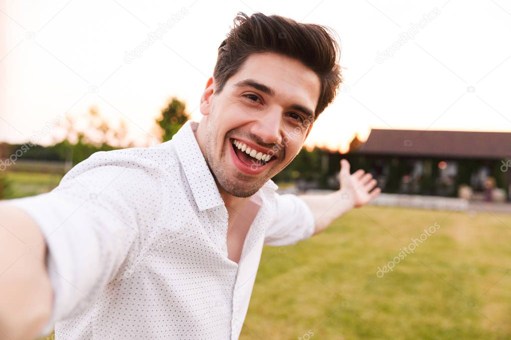 Image of happy adult man wearing white shirt laughing while taking selfie during walk on nature near country house