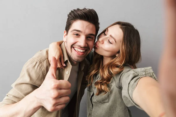 一对快乐的年轻夫妇的肖像拥抱 而采取了自拍和显示大拇指上孤立的灰色背景 — 图库照片