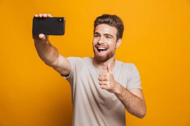 Yapmak imge-in yakışıklı mutlu adam sarı duvar backgroung üzerinde izole ayakta bir selfie yanında smartphone ile başparmak.