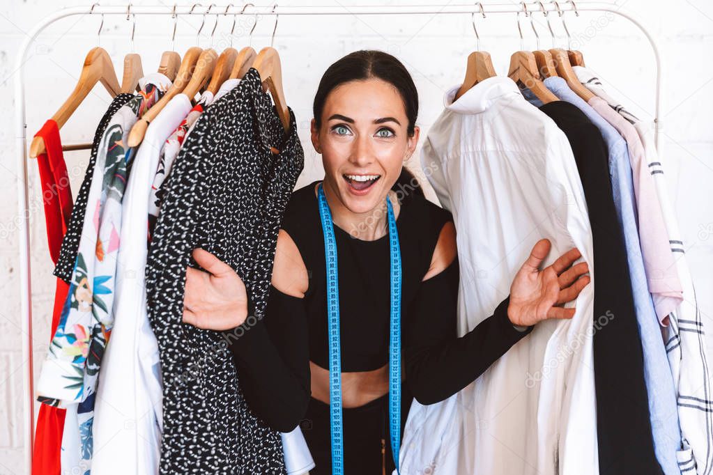 Excited young woman clothes designer standing at the clothes rack indoors