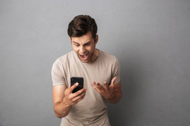 T-shirt çığlık içinde tahriş olmuş adam 20'li yaşlarda ise gri arka plan üzerinde izole smartphone bakarak öfkeyle