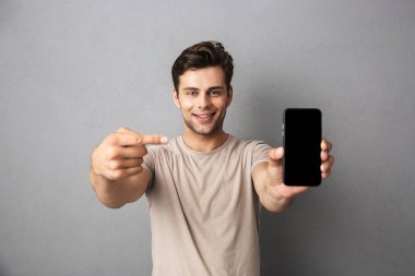 Boş ekran cep telefonu gösterilen gri artalan üzerinde izole t-shirt neşeli bir genç adam portresi