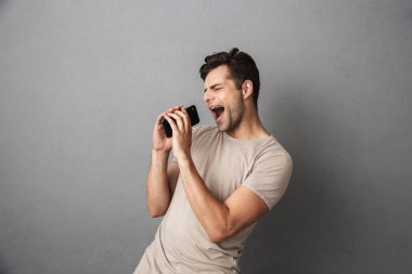Gri artalan üzerinde izole t-shirt şarkı boş ekran cep telefonu, holding neşeli bir genç adam portresi