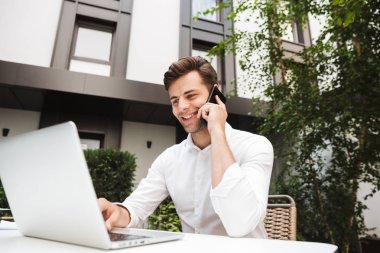 Kendine güvenen genç işadamı ile dizüstü bilgisayar açık havada otururken cep telefonu üzerinde konuşurken gömlek giymiş