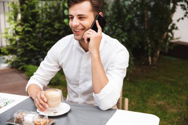 Cep telefonu üzerinde konuşurken kafe açık havada kahve içme gömlek gülümseyen genç işadamı giymiş