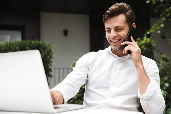 ラップトップ コンピューター屋外で座りながら携帯電話で話しているシャツを着ている幸せな若いビジネスマン — ストック写真