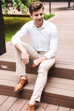 Gülümseyen genç erkek Yöneticisi resmi giyinmiş cep telefonu holding, açık havada oturma