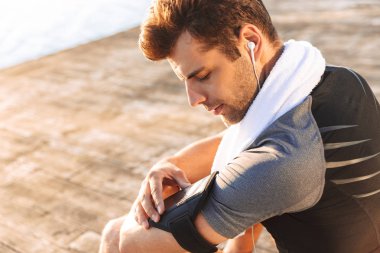 Eşofman dinleme runner adam 20'li yaşlarda smartphone kol tutucu, ahşap iskele, sahil üzerinde otururken kullanarak kablosuz kulaklık ile müzik için kırpılmış fotoğrafı