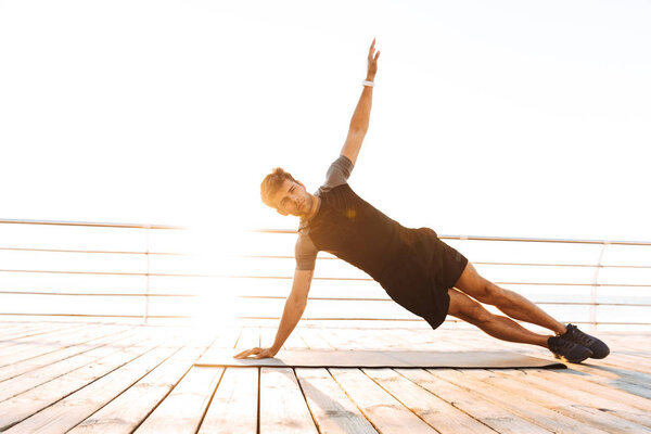 Photo of strong european man 20s in tracksuit raising arm and doing side plank on mat while working out on wooden pier at seaside in morning