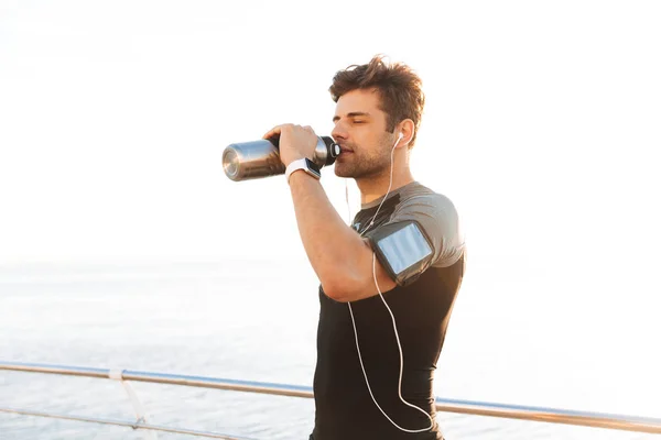 运动的欧洲人的相片20S T恤听音乐通过无线耳机和饮用水从金属杯子在海滩训练以后 — 图库照片