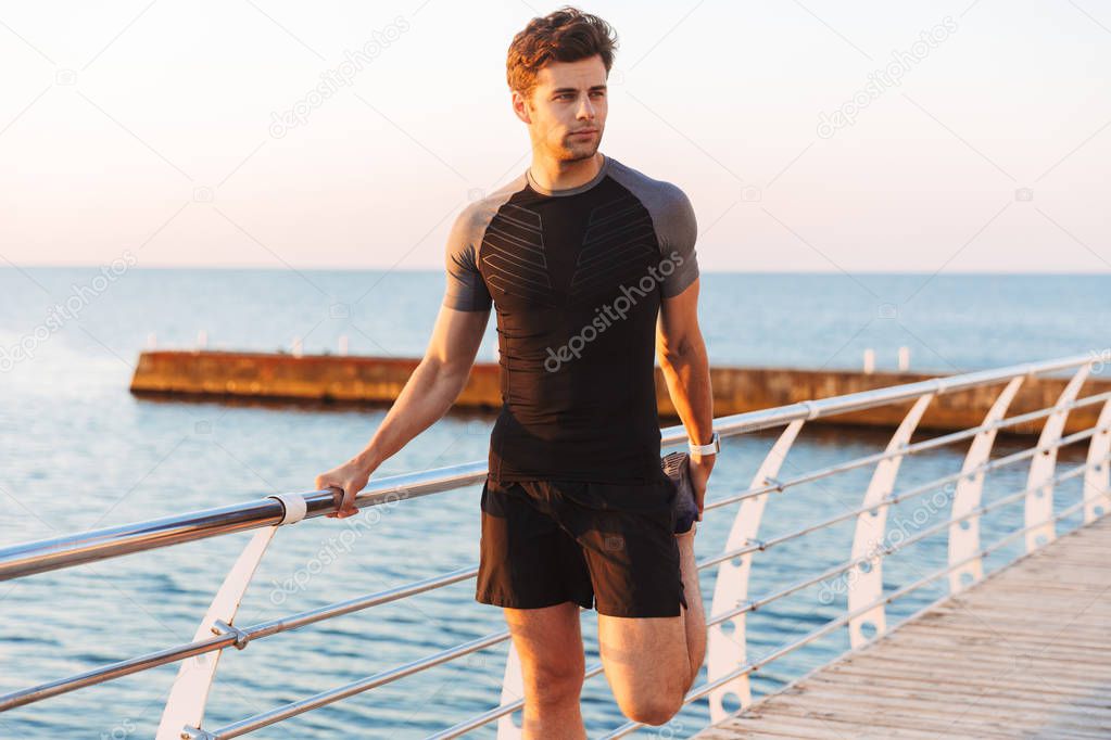 Image of muscular sporty man 20s in shorts and t-shirt working out and stretching legs on pier or boardwalk at seaside in morning