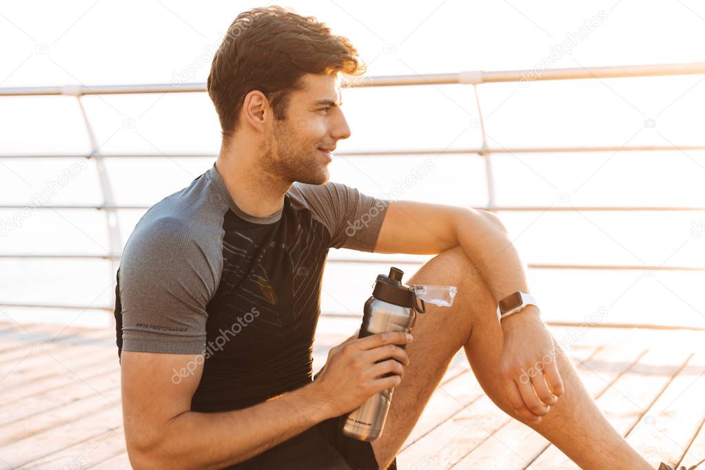 Photo of handsome sportsman 20s in tracksuit sitting on mat with thermos mug after workout on wooden pier at seaside in morning