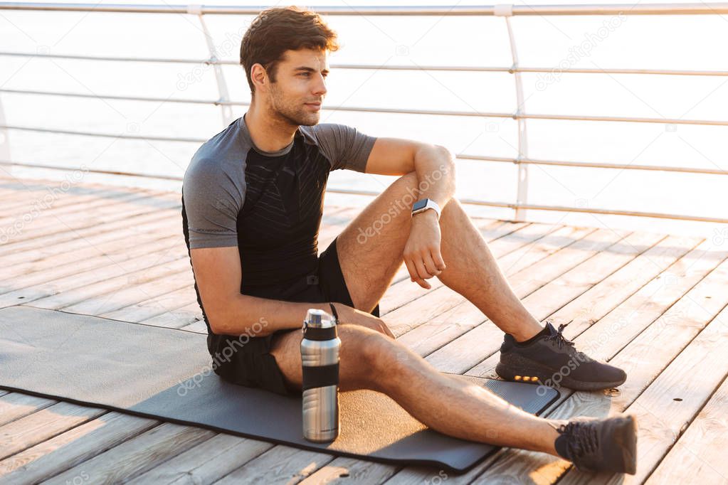 Photo of adult sportsman 20s in tracksuit sitting on mat with thermos mug after workout on wooden pier at seaside in morning