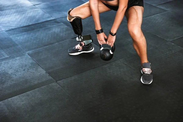 Portrait of sporty disabled woman with prosthesis in tracksuit doing crossfit exercises and lifting dumbbell in gym