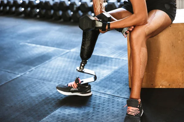 Cropped image of fitness disabled invalid woman wearing prosthesis in tracksuit training and lifting dumbbell in gym