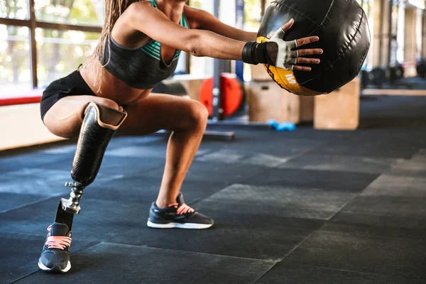 Portrait of beautiful invalid woman with prosthesis in tracksuit doing sit-ups with fitness ball in gym