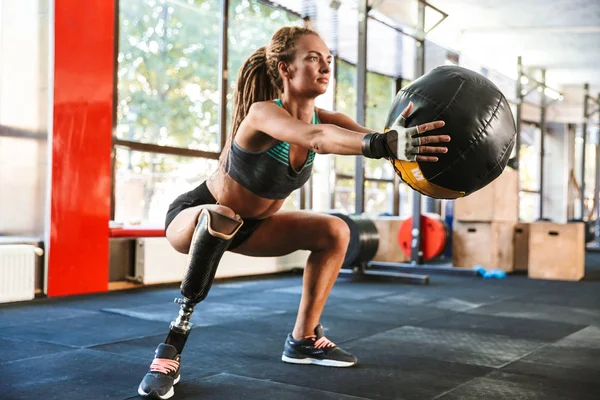 Portrait of european invalid woman with prosthesis in tracksuit doing sit-ups with fitness ball in gym