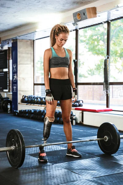 Portrait of strong caucasian disabled woman wearing prosthesis in tracksuit training and lifting barbell in gym
