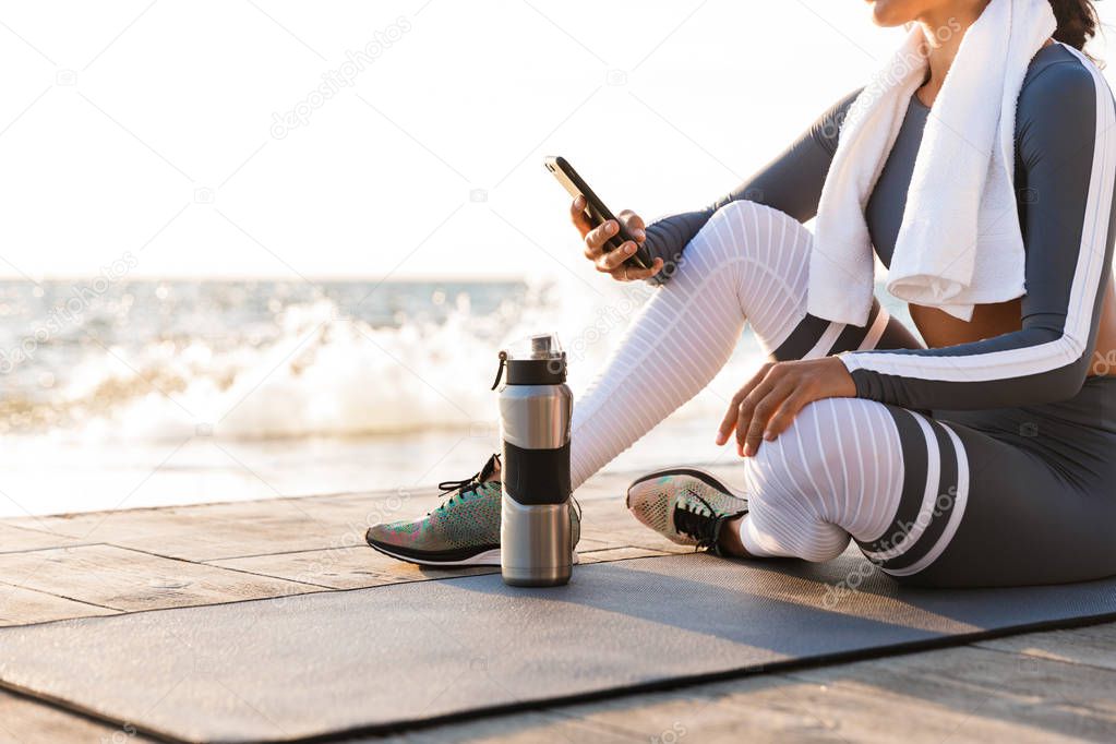 Cropped photo of young fitness woman outdoors on the beach have a rest sitting with towel and water using mobile phone.