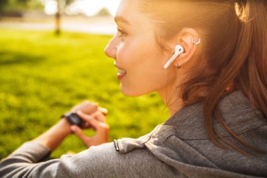 Portrait of beautiful sporty woman 20s in sportswear using smartwatch and wireless earbud while resting in green park clipart