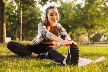 Görüntü gülümseyen fitness, egzersiz ve egzersiz üzerinde otururken bacak germe kadın 20'li yaşlarda giyen kulaklık mat yeşil park
