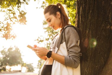 Fotoğraf gülümseyen ve yeşil parkta yürürken smartphone tutan spor giyim sportif beyaz kadının 20'li