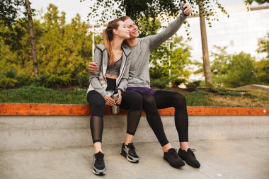 Görüntü genç sportif çift erkek ve kadın 20'li yaşlarda egzersiz veya koşu gün güneşli yaz sonra yeşil park ve alma selfie parapetin üzerinde oturan eşofman