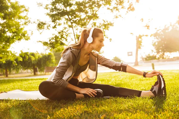 微笑运动员20S 戴着耳机工作和伸展腿坐在绿色公园的运动垫上的形象 — 图库照片